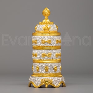 May include: A decorative, ornate container with a silver and gold finish. The container features a tiered design with gold accents, including bows and a crown-like top. The base has a detailed, curved design.