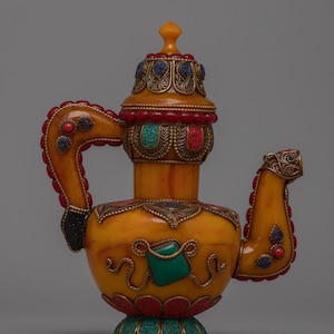 Könnte beinhalten: Verzierte gelbe Teekanne mit aufwendigen Metallarbeiten und farbigen Edelsteinakzenten. Die Teekanne hat einen gebogenen Griff und Ausguss, einen dekorativen Deckel und einen Sockel mit floralem Design. Die Edelsteine sind rot, blau und grün.