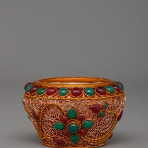 May include: An ornate bowl with a copper filigree design. The bowl is decorated with red and green gemstones around the rim and on the body. The bowl has a gold interior and a rounded shape. The bowl's measurements are in centimetres.