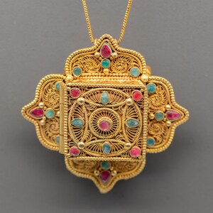 Könnte beinhalten: Eine goldfarbene Anhänger-Halskette mit filigranem Design. Der Anhänger ist mit roten und türkisfarbenen Edelsteinen verziert. Die Halskette hat ein aufwendiges, durchbrochenes Design mit einem Scharnierfach.
