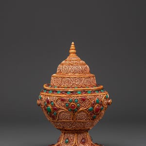 May include: Ornate gold-colored decorative vessel with a lid, adorned with intricate filigree and inlaid with red and green gemstones. The vessel has a pedestal base and a conical top, showcasing detailed craftsmanship.
