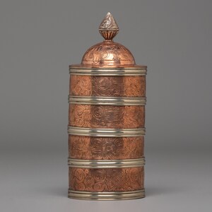 Puede incluir: Un recipiente de color cobre con adornos y tapa plateados. El recipiente tiene forma cilíndrica con un motivo floral decorativo. La tapa es abovedada y rematada con un adorno plateado. El recipiente probablemente se utiliza para el almacenamiento.