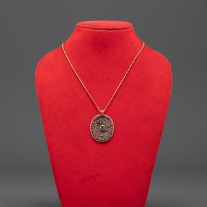 Può includere: Una collana con pendente color oro con un design ovale a filigrana. Il pendente presenta una pietra verde centrale circondata da pietre turchesi e rosse più piccole. La collana è esposta su un busto rosso su sfondo grigio.