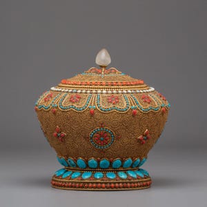 Peut inclure: Un récipient décoratif doré orné d'un couvercle, orné de pierres turquoise, rouges et blanches. Le récipient présente des motifs de filigrane complexes et une pierre transparente en forme de goutte d'eau sur le dessus. La base est ornée de pierres turquoises.