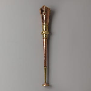 May include: A copper and brass ceremonial staff with a red gemstone. The top section is a copper-colored, shield-shaped design with a gold trim. The staff has a gold band and a long, copper-colored shaft, transitioning to a gold base.