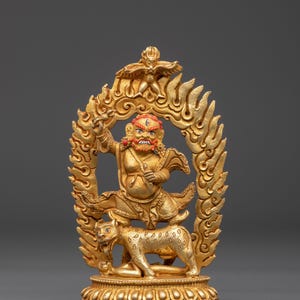 May include: A golden statue of a deity with a fierce expression, red beard, and a flaming halo. The deity stands above a golden tiger. The statue is intricately detailed and sits on a decorative base.