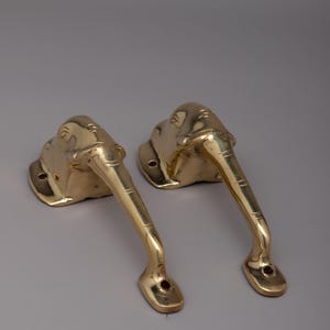 May include: Two brass elephant-shaped door handles. The handles are gold-colored and feature detailed elephant head designs. The handles have a curved, elongated shape with a hole at the base for mounting. The handles are likely for a door or cabinet.