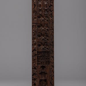 Könnte beinhalten: Ein dunkelbraunes, rechteckiges Holzschnitzwerk mit aufwendigen Reliefdetails. Die Schnitzerei zeigt verschiedene florale, geometrische und menschliche Figuren. Das Design ist vertikal ausgerichtet, mit einem detaillierten Muster über die gesamte Länge des Holzes.