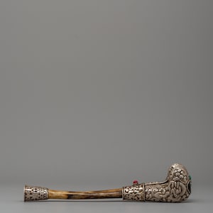 May include: An ornate, antique pipe with a curved stem. The pipe features silver-colored metal detailing with intricate floral patterns and is adorned with red and turquoise gemstones. The stem is a light brown color.