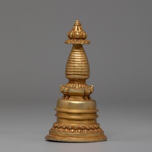 May include: A gold-coloured, ornate stupa with a tiered base and a conical top. The stupa features detailed carvings and a ribbed, cylindrical body. The design includes lotus-shaped elements and a rounded base with a beaded border, showcasing a traditional religious artifact.