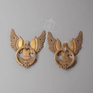 Könnte beinhalten: Ein Paar Türklopfer aus Messing, die jeweils einen Vogel mit ausgebreiteten Flügeln und einem in der Mitte gehaltenen kreisförmigen Ring zeigen. Das Metall hat einen warmen Goldton mit detaillierten Federgravuren und einem Blumenakzent.