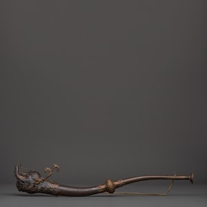May include: A long, dark brown ceremonial horn with ornate brass detailing. The horn features a carved dragon head at the mouthpiece and a flared bell at the end. A brass cord is attached to the horn.