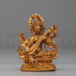 Könnte beinhalten: Eine goldfarbene Metallstatue einer Gottheit, die auf einem Lotussockel sitzt. Die Figur hat vier Arme, trägt eine verzierte Krone sowie Schmuck und spielt ein Saiteninstrument. Die Skulptur weist detaillierte Schnitzereien und Muster auf.