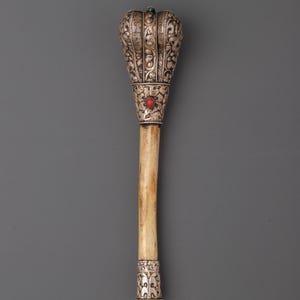 May include: An ornate ceremonial mace with a bone shaft and silver-colored metalwork. The top is elaborately decorated with floral patterns and set with a red and green gemstone. The mace is a symbol of authority and power.