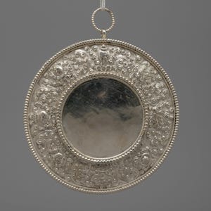 May include: A silver-colored, circular pendant with intricate floral and scrollwork designs. The pendant has a central, reflective disc and a beaded border. A small ring is attached at the top, connected to a thin, white string for hanging.