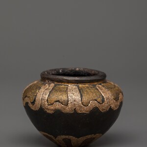 Könnte beinhalten: Ein kleiner, runder, dekorativer Topf mit dunkelbraunem Boden und einem goldfarbenen, wellenförmigen Design am oberen Rand. Der Rand ist ebenfalls dunkelbraun. Der Topf hat ein rustikales, handwerkliches Aussehen.