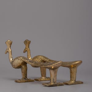 May include: Three brass peacock-shaped decorative objects. Each has a detailed body, a long, curved tail, and a small base. The objects are golden in color and stand on a gray background.