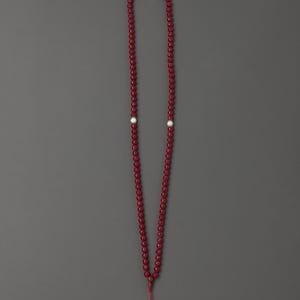 以下が含まれることがあります： 3つの白いビーズと赤いタッセルが付いた、長い深紅色のビーズネックレス。ビーズは小さくて丸く、ネックレスは灰色の背景に表示されています。