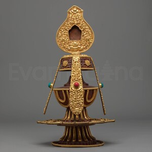 Peut inclure: Un objet décoratif doré orné, avec un sommet en forme de goutte d'eau et une base à plusieurs niveaux. L'objet présente des sculptures complexes, une pierre précieuse rouge et deux pierres précieuses turquoise. Le design est symétrique et détaillé.