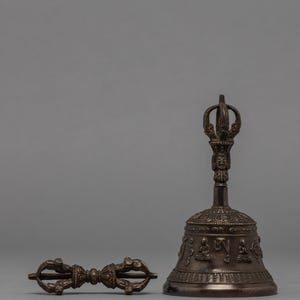 May include: A bronze-colored Buddhist bell and a dorje, a ritual object. The bell has a detailed handle and is decorated with figures and inscriptions. The dorje is a symmetrical, hand-held object with intricate designs. Both items are on a gray background.