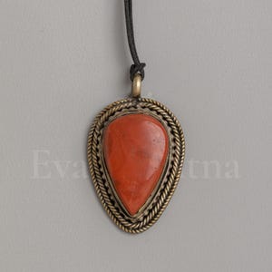 May include: A teardrop-shaped pendant featuring a smooth, solid red stone set in a brass-toned metal frame with a twisted rope border. The pendant hangs from a thin black cord and includes a small metal loop at the top for the cord attachment.