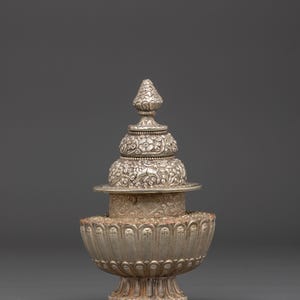 May include: A silver-colored, ornate decorative object with a tiered design. The base is a bowl shape with a floral pattern, supporting a stack of detailed, engraved sections, topped with a conical finial. The object has an aged, antique appearance.
