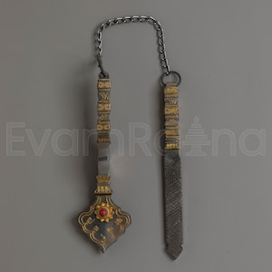 May include: An ornate, antique metal object with a chain. The object has a decorative handle with gold accents and a red stone. The other end has a textured, dark grey blade-like shape. The chain is silver.