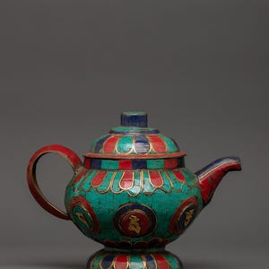 May include: A decorative teapot featuring a mosaic of inlaid turquoise, red, and blue stones set in metal. The body and lid are adorned with repeating petal-like patterns and circular medallions containing gold-colored symbols on a dark background.
