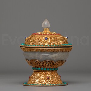 Peut inclure: Un récipient doré orné d'un couvercle, orné de pierres précieuses turquoise, rouges et bleues. Le récipient a un corps arrondi gris clair et un fleuron clair et pointu sur le dessus. Le design complexe suggère un but cérémoniel ou décoratif.