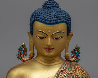 Puede incluir: Busto de Buda dorado con cabeza azul y detalles intrincados. El busto presenta una expresión serena, labios rojos y adornos decorativos en los hombros con detalles en rojo, verde y azul. Es un objeto religioso.