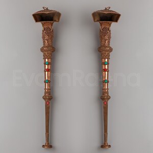 May include: Two ornate, bronze-colored ceremonial staffs. Each staff features a flared top with intricate carvings, a central section adorned with red and turquoise stones, and a tapered lower portion. The staffs are displayed against a neutral gray background.