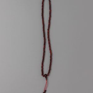 Pode incluir: Um colar longo de contas castanho-avermelhadas com uma conta central maior e uma borla vermelha. O colar é feito de pequenas contas redondas e é exibido em um fundo cinza. A borla tem algumas contas menores na extremidade.