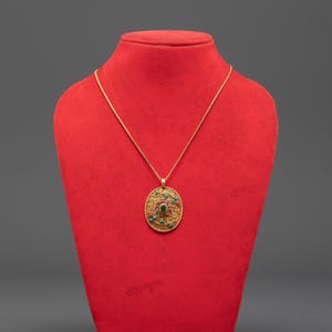 May include: A gold-colored pendant necklace with an oval-shaped pendant. The pendant is adorned with red, green, and turquoise-colored stones. The necklace is displayed on a red bust against a gray background.