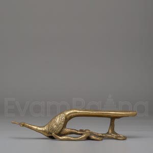 May include: A brass-colored decorative object in the shape of a bird, possibly a peacock or crane. The bird is lying down with its head and tail extended. The object has a flat, rectangular surface supported by a single leg.