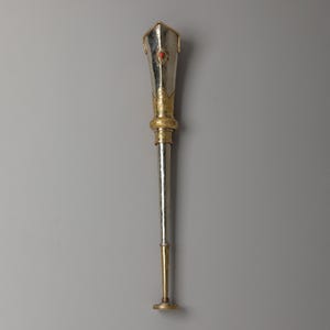 May include: A ceremonial staff with a silver shaft and a gold-colored top and base. The top features a red gemstone and gold accents. The staff is approximately 24 inches long.