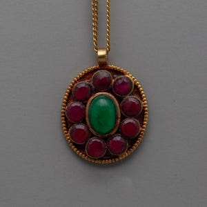 May include: A gold pendant necklace featuring a central oval green gemstone surrounded by ten round red gemstones. The pendant has a textured gold border and hangs from a delicate gold chain.