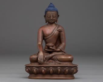 May include: A bronze-colored Buddha statue with a blue head covering. The seated figure holds a small bowl and is positioned on a detailed, ornate base. The statue has a serene expression and is a representation of Buddhist art.