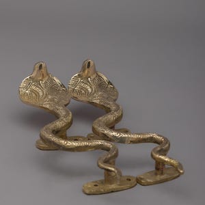 May include: Two brass door handles in the shape of cobras. The handles are gold-colored with detailed scale patterns and are mounted on rectangular bases. The cobra heads are raised and detailed.