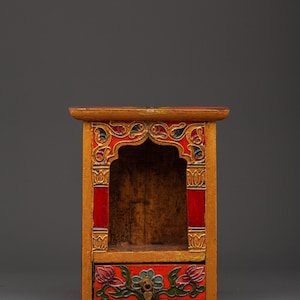May include: A small, ornate wooden cabinet with a drawer. The cabinet is painted in gold, red, and blue, with floral and geometric designs. The drawer features a floral design and a small metal handle. The cabinet has a small arched opening.