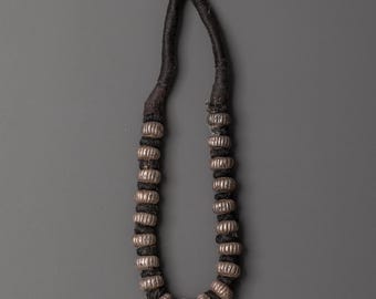 May include: A handcrafted necklace featuring silver-colored, ribbed beads strung on a dark, braided cord. The necklace has a simple, elegant design, with the beads evenly spaced along the cord. The cord is knotted at the top.