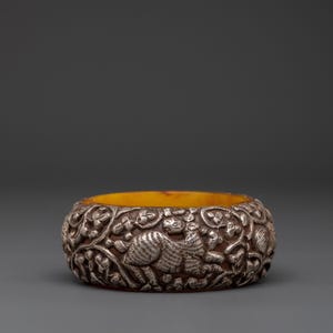 Puede incluir: Un brazalete ancho de color plateado con un intrincado diseño en relieve. La detallada talla representa animales y follaje. El interior del brazalete es de color amarillo cálido. El brazalete es una pieza llamativa.
