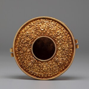 May include: A gold-colored, circular container with intricate floral and scrollwork patterns. The container has a central opening and is adorned with detailed embellishments. Two small clasps are visible on the sides. The overall design suggests a decorative or ceremonial object.