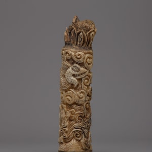 May include: A carved, off-white cylindrical sculpture with intricate details. The sculpture features a dragon-like creature amidst swirling clouds and decorative patterns. The top has a textured, organic shape.