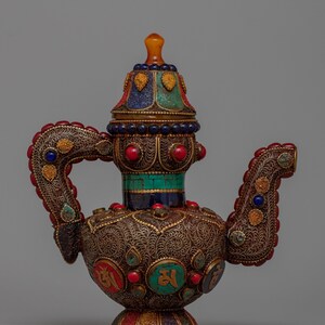 Può includere: Teiera antica ornata con intricati lavori in filigrana metallica. La teiera presenta una varietà di pietre preziose colorate, tra cui rosso, blu e turchese, e un coperchio decorativo. Il manico e il beccuccio sono anch'essi adornati con pietre preziose. Il design è dettagliato e artistico.