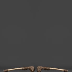 May include: Two antique pipes with ornate, detailed carvings on the bowl and stem ends. The bowls are a dark bronze color, and the stems are a light beige. The pipes are set against a solid gray background.
