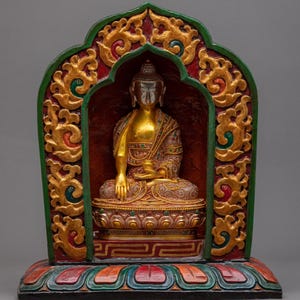 Op de afbeelding: Een gouden Boeddha-beeld in een meditatieve houding, gehuisvest in een decoratief, sierlijk houten frame. Het frame heeft rode, groene en gouden accenten met ingewikkelde gravures. De basis is beschilderd met kleurrijke patronen.