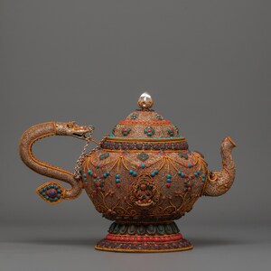 Peut inclure: Théière antique ornée avec un travail de métal complexe. La théière présente une poignée en forme de dragon, un bec verseur et est ornée d'accents turquoise et rouges. Le couvercle est surmonté d'un bouton en forme de gemme transparente. La base est décorée de motifs rouges et dorés.