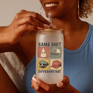 May include: Frosted glass tumbler with a bamboo lid. The tumbler features the text "SAME SHIT DIFFERENT HAT" with four images of different hats. The tumbler is held by a person wearing a blue tank top.