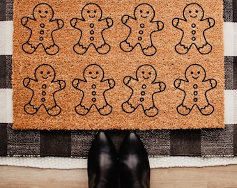 May include: A rectangular doormat with a light brown coir surface features eight black gingerbread man outlines. The mat is placed on a black and white checkered rug, with a person's black boots and blue jeans visible.