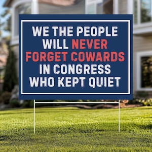 May include: A blue yard sign with white and red text that reads "WE THE PEOPLE WILL NEVER FORGET COWARDS IN CONGRESS WHO KEPT QUIET." The sign is in a white frame and is held up by two metal stakes in a grassy yard.